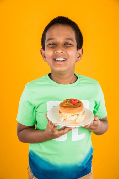 Söpö intialainen poika ja hampurilainen pieni valkoinen levy, pieni aasialainen poika ja hampurilainen, yli värikäs tausta — kuvapankkivalokuva