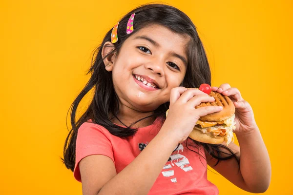 Bonito indiana menina comer hambúrguer, pequeno asiático menina e hambúrguer, isolado sobre amarelo fundo — Fotografia de Stock