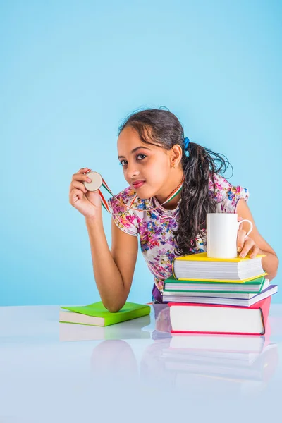 Chica estudiosa india mostrando medalla de oro o signo de éxito, inclinándose sobre la mesa con pila de libros, niña asiática y concepto de éxito, 1º en concepto de estudios, colegiala india y concepto de mérito — Foto de Stock