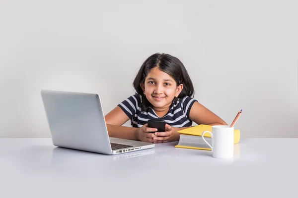 Cute little indian girl using smartphone while studying on laptop — Stock Photo, Image