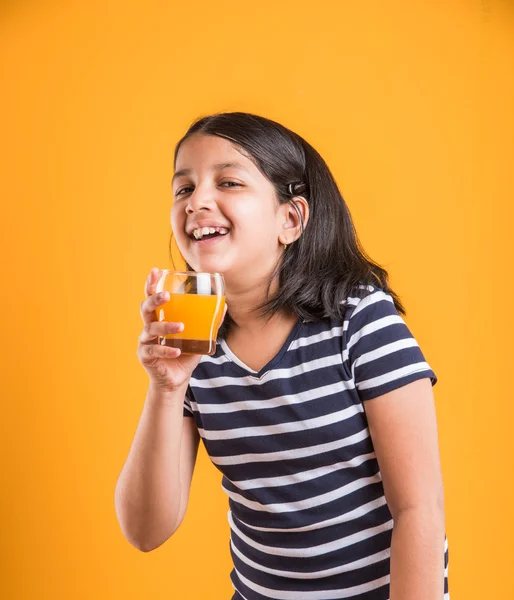 portrait of indian small girl drinking mango juice or fruit juice in a glass, asian girl and a glass of juice, indian small girl holding a glass of mango juice or orange juice on yellow background