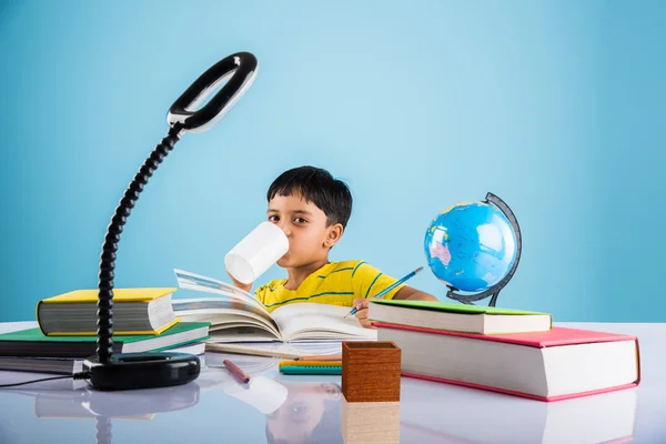 Индийский маленький мальчик, изучающий или выполняющий домашнюю работу, азиатский мальчик, занимающийся с кофейной кружкой, моделью глобуса и книгами на столе — стоковое фото