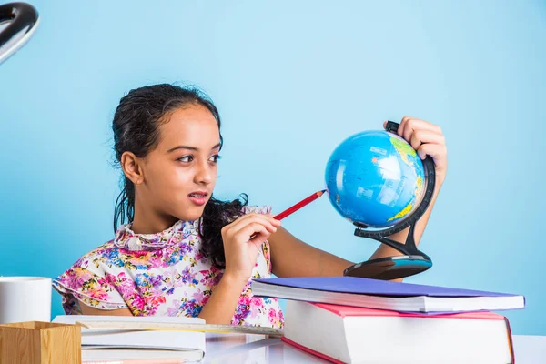 indian girl student looking at educational globe and sitting at table with books, table lamp and milk mug, asian girl child studying geography, curious asian girl studying geography with globe