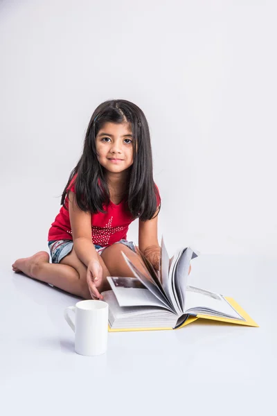 Індійська маленька дівчинка читає книгу, азіатська дівчинка читає книгу на білому тлі, мила 5 річна індійська маленька дівчинка читає книгу з молочним кухоль — стокове фото