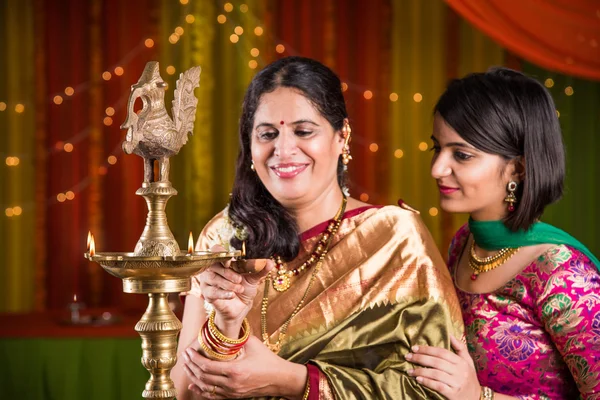 Indian young girl in traditional wear and mother in saree lighting oil lamp or samai with diya and celebrating ganesh festival or Diwali or deepavali. Indian lady hands holding oil lamp indoors.