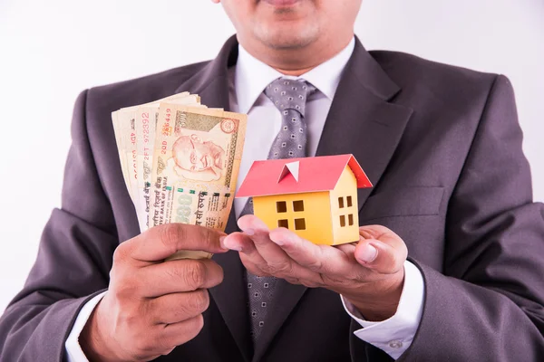 Zakup koncepcji domu, indyjski mężczyzna gospodarstwa indyjskich notatek walutowych w jednej ręce i mały dom model w drugiej ręce, zbliżenie i selektywne skupienie — Zdjęcie stockowe