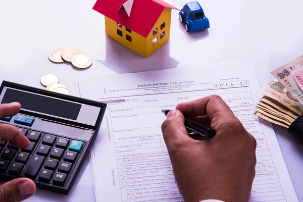 Hombre indio o contable persona que presenta formulario de declaración de impuestos indio o documento ITR que muestra moneda india, modelo de casa, coche de juguete y calculadora sobre mesa blanca, enfoque selectivo —  Fotos de Stock