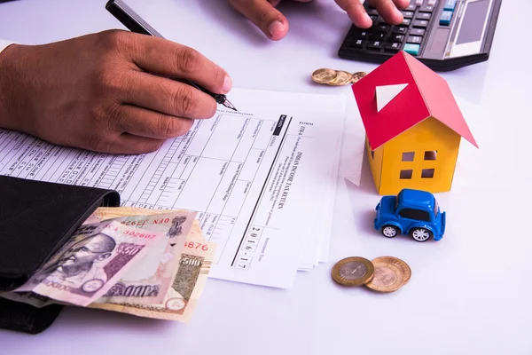 Hombre indio o contable persona que presenta formulario de declaración de impuestos indio o documento ITR que muestra moneda india, modelo de casa, coche de juguete y calculadora sobre mesa blanca, enfoque selectivo —  Fotos de Stock