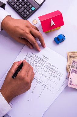 Indian man or accountant person filing Indian income tax returns form or ITR document showing indian currency, house model, toy car and calculator over white table top, selective focus clipart