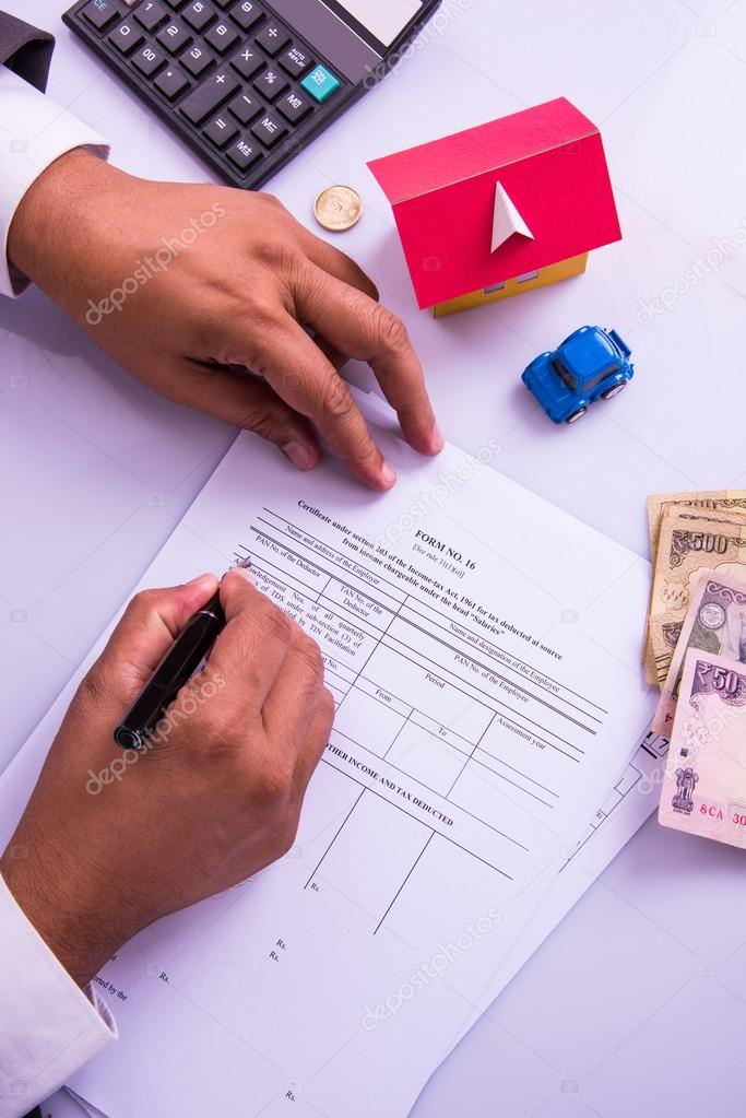 Indian man or accountant person filing Indian income tax returns form or ITR document showing indian currency, house model, toy car and calculator over white table top, selective focus
