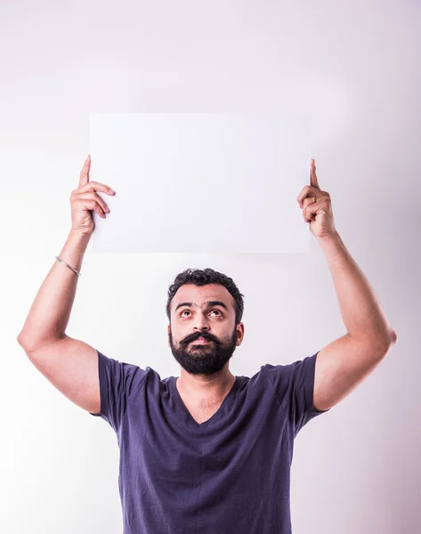 Retrato de homem indiano bonito com barba segurando placa branca em branco. homem indiano segurando cartaz em branco . — Fotografia de Stock