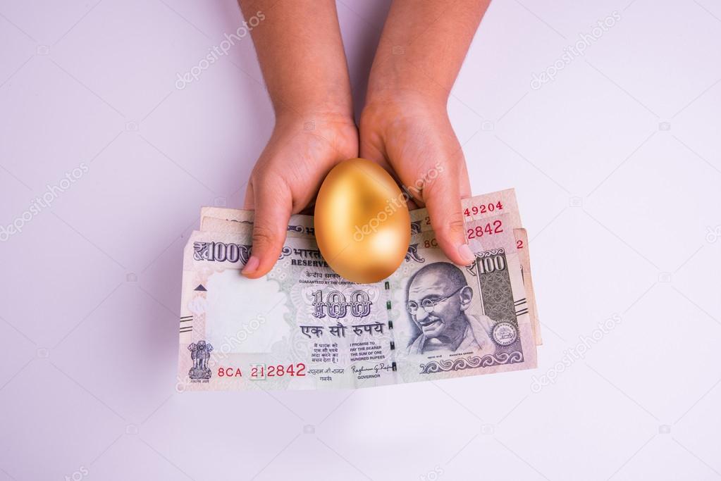 closeup of small girl holding indian currency notes or hundred rupee notes and golden egg over it