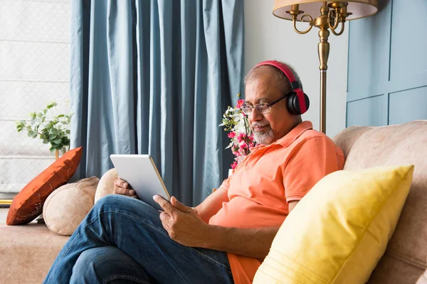 Indian asian old man using headphones while listning to music or watching movies on tablet computer or smartphones