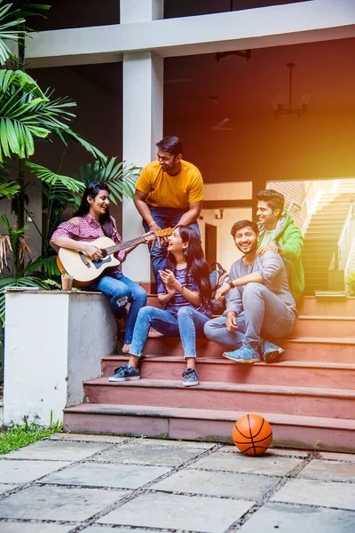 Asian Indian college students playing music with guitar while sitting in campus on stairs or over lawn