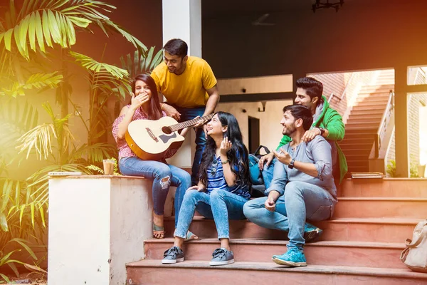 Asian Indian college students playing music with guitar while sitting in campus on stairs or over lawn