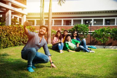 Asyalı Hintli üniversite öğrencileri kampüste merdivenlerde ya da çimlerde oturup eğlenirken akıllı telefon kullanarak selfie çekiyorlar.