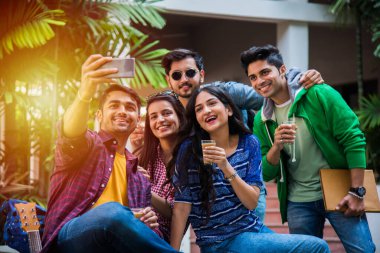 Asyalı Hintli üniversite öğrencileri kampüste merdivenlerde ya da çimlerde oturup eğlenirken akıllı telefon kullanarak selfie çekiyorlar.