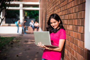 Asyalı Hintli üniversite öğrencisi dizüstü bilgisayar ya da kitap üzerinde çalışırken diğer sınıf arkadaşları arka planda üniversite kampüsünde açık havada resim çekiyor.