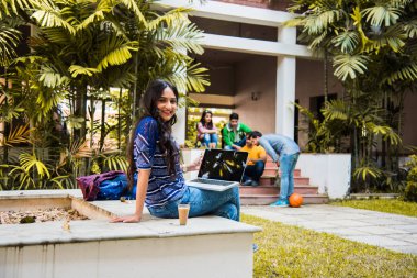 Asyalı Hintli üniversite öğrencisi dizüstü bilgisayar ya da kitap üzerinde çalışırken diğer sınıf arkadaşları arka planda üniversite kampüsünde açık havada resim çekiyor.