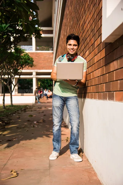 Asian Indian college student in focus working on laptop or reading book while other classmates in the background, outdoor picture in university campus