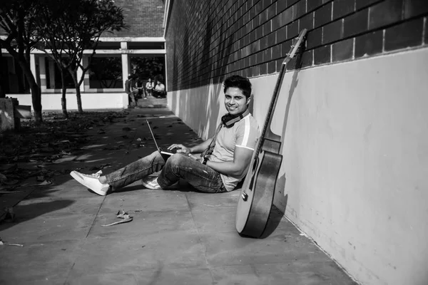 Asian Indian college student in focus working on laptop or reading book while other classmates in the background, outdoor picture in university campus