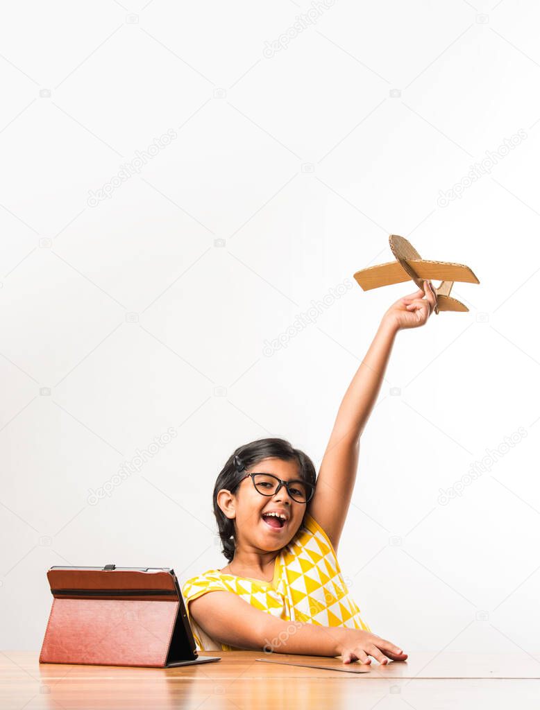 Indian small girl student making or measuring wings of paper plane or Aeroplane, doing school project or learning science with online tutorial