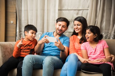 Hintli aile kanepede oturup akıllı telefon, dizüstü bilgisayar ya da tablet kullanıyor, film izliyor ya da internette sörf yapıyor.