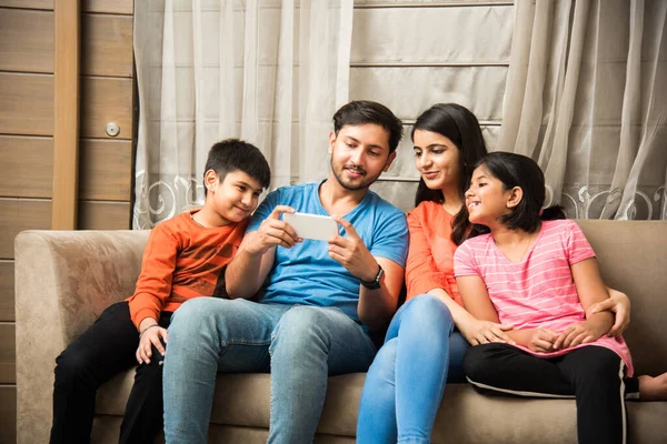 Indian family sitting on sofa and using smartphone, laptop or tablet, watching movie or surfing internet
