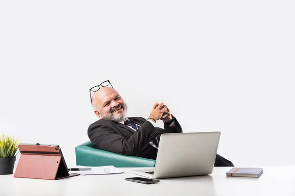 コンピュータの前の職場に座って リラックスしながらカメラを見ている満足したインドのシニアアジアのビジネスマンの肖像画 オフィスの机の上に古いプロの男の足 — ストック写真