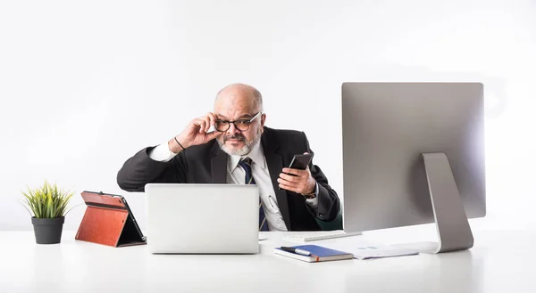 アジア系インド人のシニア金融ビジネスマンは 彼のワークステーションや机にコンピュータ ラップトップ タブレットの前に座っています 書類を書いている間に電話で話す — ストック写真