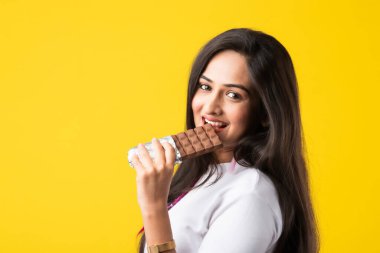 Pembe ya da sarı arka planda izole edilmiş çikolata yiyen mutlu bir Hintli genç kadının portresi.