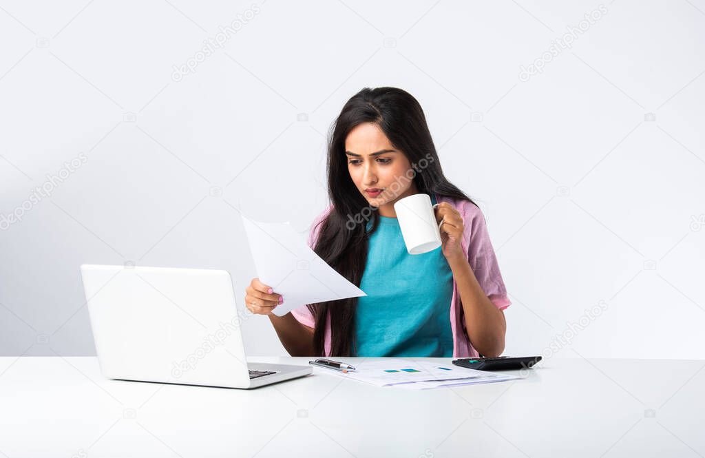 Serious focused Indian businesswoman or house wife holding papers preparing report ,analyzing work results, doing paperwork at workplace using computer online software