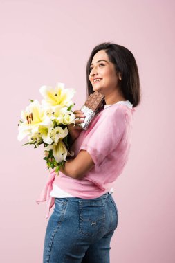 Çiçek buketi ve çikolata tutan genç ve çekici bir Hintli kadının portresi. Pembe arka planda izole edilmiş duruyor