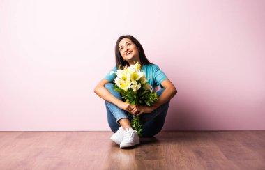 Ahşap zeminde pembe duvara yaslanmış bir şekilde otururken elinde çiçek buketi tutan çekici bir Hintli kadının portresini yakından çek.