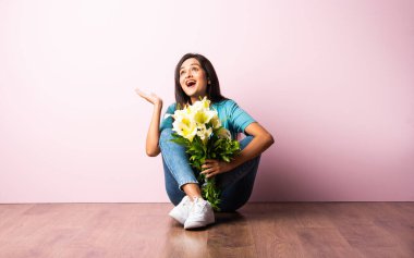 Ahşap zeminde pembe duvara yaslanmış bir şekilde otururken elinde çiçek buketi tutan çekici bir Hintli kadının portresini yakından çek.