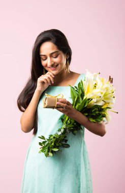 Çekici genç bir Hintli kadının portresi. Elinde çiçek buketi tutarken hediye kutusunu açıyor. Pembe arka planda tek başına duruyor.
