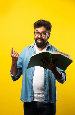 Genç Hintli mutlu adam gözlük takıyor kitap okuyor ve sarı arka planda izole bir şekilde gülümsüyor.