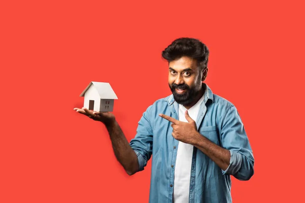 Real estate buying and people concept - Indian bearded man with miniature house model, keys and piggy bank against red background
