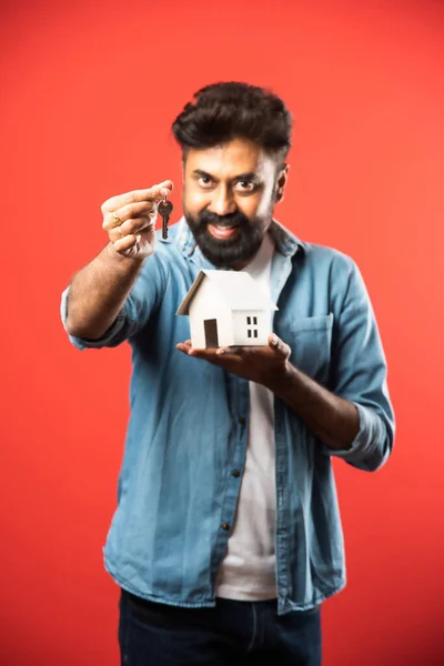 Real estate buying and people concept - Indian bearded man with miniature house model, keys and piggy bank against red background