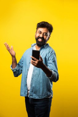 Genç sakallı Hintli adam akıllı telefon kullanıyor, ararken ya da sohbet ederken gülümsüyor, sarı arka planda duruyor.