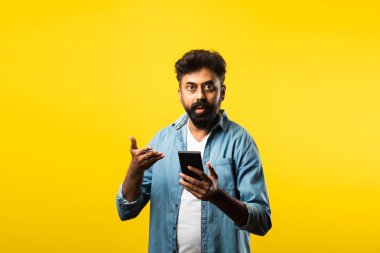 Genç sakallı Hintli adam akıllı telefon kullanıyor, ararken ya da sohbet ederken gülümsüyor, sarı arka planda duruyor.