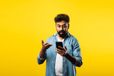 Genç sakallı Hintli adam akıllı telefon kullanıyor, ararken ya da sohbet ederken gülümsüyor, sarı arka planda duruyor.