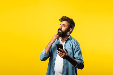 Genç sakallı Hintli adam akıllı telefon kullanıyor, ararken ya da sohbet ederken gülümsüyor, sarı arka planda duruyor.