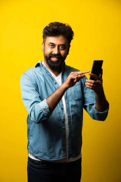 Young bearded Indian man using smartphone, smiling while calling or chatting with friend, standing against yellow background