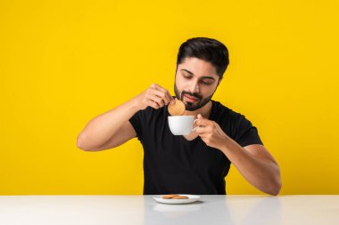 Yakışıklı, sakallı, sağlıklı tahıllı bisküvi yiyen çay fincanına batırılmış, sarı arka planda masada oturan Hintli bir adam.