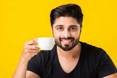 Hintli genç adam elinde çay fincanı tutuyor, içiyor ve kahvenin tadını çıkarıyor. Asyalı adam sarı arka planda, masada oturuyor.