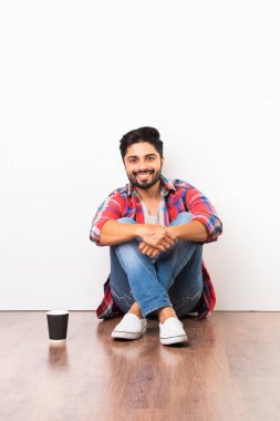 Kızılderili sakallı genç adam, tek kullanımlık kağıt bardaktan kahve içerken beyaz arka planda, ahşap zeminde izole bir şekilde oturuyor.