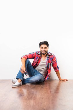 Asyalı mutlu bir adamın portresi ahşap zeminde beyaz duvarın arkasında kameraya bakarak tek başına oturuyor.