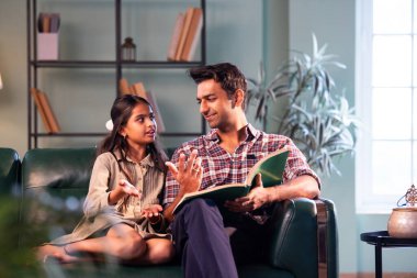 Kızına kitap okuyor, Hintli baba ve kızı modern evdeki koltukta oturuyor, Asyalı baba genç kızla bağ kuruyor, hikayeyi paylaşıyor, aile kalitesi, ev içi öğrenme ve duyguları paylaşıyor.