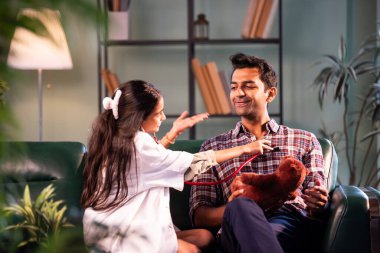 Oyuncak ayı ve babanın kalp atışlarını kontrol ediyorum, doktor gibi giyinmiş Hintli kız babasını inceliyor, ikisi de modern evdeki koltukta oturuyor, Asyalı kız ailesiyle bağ kuruyor.